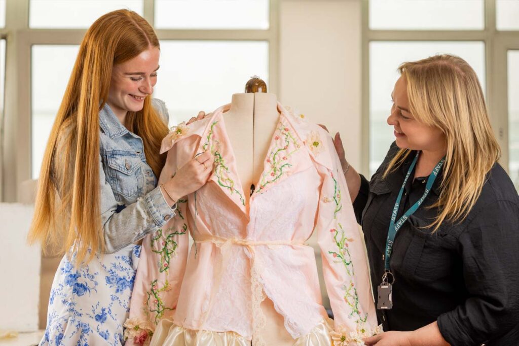 Student stitching embroidery on a dress with a tutor directing her