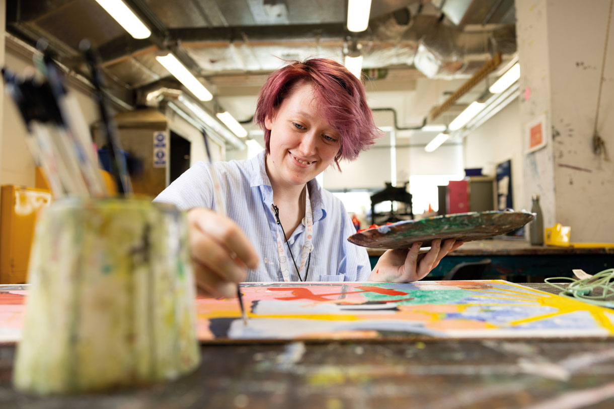 Student painting a canvas while holding a paint palette